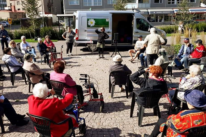 "Useita vanhuksia istuu tuoleissa puistossa. Monella on rollaattori mukana. Puistotoiminnan vetäjät ovat pakettiauton edessä."